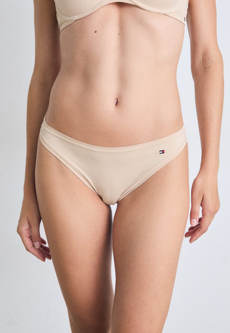 Femeie îmbrăcată în bikini și sutien bej, stând cu brațele relaxate pe lângă corp, pe un fundal simplu.