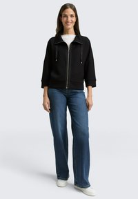 Zwarte jas met ritssluiting en een geribbelde textuur met twee trekkoorden, gecombineerd met lichtblauwe wijduitlopende spijkerbroek en witte sneakers.