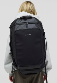 Czarny plecak o strukturalnym kształcie, wykonany z wytrzymałego materiału. Posiada główną komorę na zamek błyskawiczny oraz logo marki z przodu.