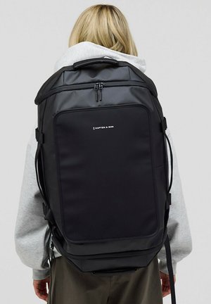 Zaino nero con una forma strutturata, realizzato in materiale resistente. Presenta un compartimento principale con zip e il logo del marchio sulla parte anteriore.