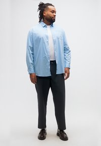 Άνδρας με dreadlocks που φοράει ένα ανοιχτό μπλε πουκάμισο πάνω από ένα λευκό μπλουζάκι, μαύρο παντελόνι και σκούρα loafers, στέκεται μπροστά σε μια απλή φόντο.