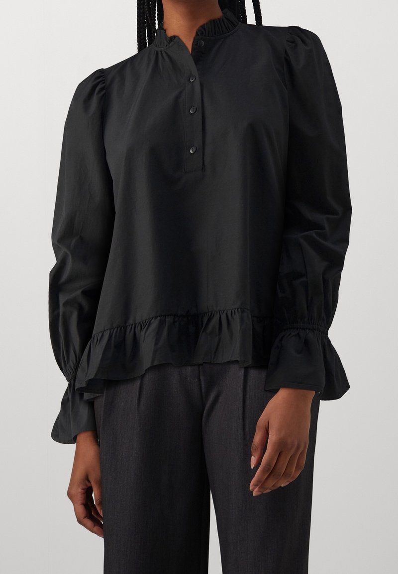 Donna che indossa una blusa nera a maniche lunghe con polsini e orlo a balze, con un bottone sul davanti, abbinata a pantaloni grigio scuro.