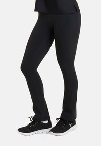 Leggings neri realizzati in tessuto elastico con una superficie liscia, design aderente e cuciture. Abbinati a scarpe sportive nere con suole bianche.