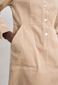 Beige Cord-Stoff Kleidungsstück mit vertikalen Rippungen, zwei Vorderaschen, sechs Metall-Druckknöpfen und einem Kragen. Betonte Nähte.