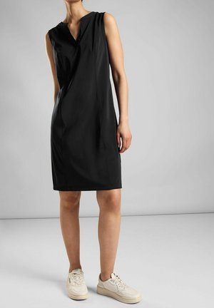 Frau trägt ärmelloses, knielanges schwarzes Kleid und weiße Sneakers und steht vor einem einfarbigen grauen Hintergrund.