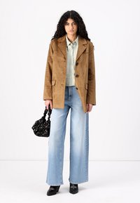 Brown suede blazer over a light green shirt; wide-leg light blue jeans; black patterned handbag; black shoes.