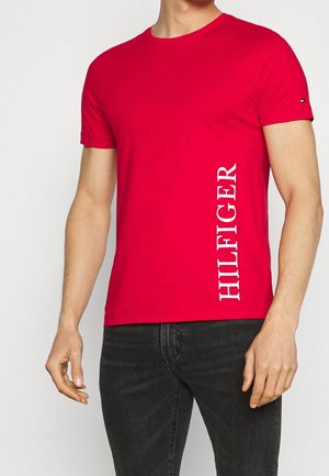 Mężczyzna noszący jasnoczerwoną koszulkę z krótkim rękawem z pionowym białym napisem „HILFIGER” po prawej stronie, połączoną z ciemnymi jeansami.