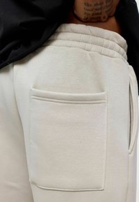 Beige trainingsbroek heeft een grote achterzak, een elastische tailleband en een zachte, textuurstof. Zichtbare stiksels voegen detail toe aan het ontwerp.