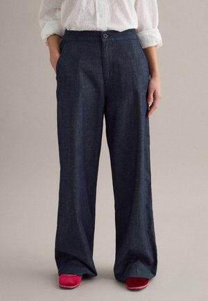 Persona con pantaloni a gamba larga blu scuro, camicia bianca a maniche lunghe con trama e scarpe rosa acceso, in piedi con le mani nelle tasche.