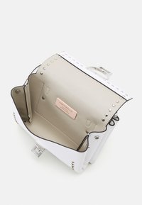 Hvid læderhåndtaske med sølvbeslag, der har en tekstureret yderside, beige inderside, dekorative nitter langs kanterne og en etiket indeni.