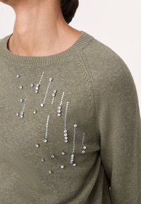 Maglione verde oliva realizzato in morbido tessuto a maglia, con un motivo irregolare di strass argentati e trasparenti su tutto il petto.