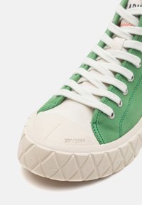 Sneaker verde in tela con suola in gomma bianca, suola testurizzata e lacci bianchi piatti. Presenta un design cucito e una linguetta con marchio.