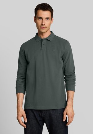 Junger Mann mit kurzen dunklen Haaren, der ein dunkelgrünes Langarm-Poloshirt und dunkle Jeans vor grauem Hintergrund trägt.