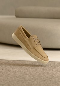 Chaussure en daim beige à lacets flottant au-dessus d'un tapis texturé marron clair avec un canapé beige flou en arrière-plan.
