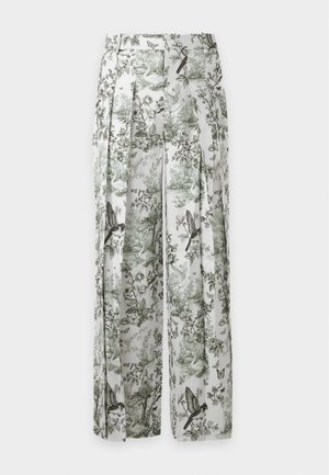 Femme portant un débardeur blanc et un pantalon blanc à jambes larges avec un imprimé nature verte et oiseaux, debout avec une main dans la poche.