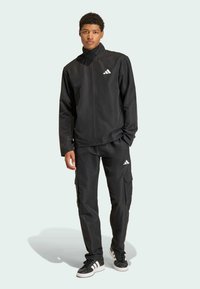 Μαύρο αθλητικό κοστούμι φτιαγμένο από λείο ύφασμα με ζακέτα με πλήρη φερμουάρ και παντελόνια cargo, με λευκές λεπτομέρειες του λογότυπου Adidas.