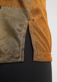 Camiseta de tela texturizada en color marrón dorado, con patrones geométricos y aberturas laterales, combinada con una prenda negra debajo.