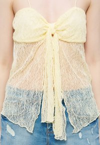 Haut sans manches en dentelle jaune avec détail de nœud devant porté sur un short en jean déchiré bleu sur une personne.