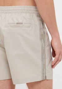 Bežové plavky so sťahovacím pásom, bočný pruh s nápisom "Calvin Klein" a zadná vrecko so klopou.