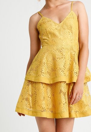 Nainen pukeutuneena hihattomaan keltaiseen pitsimekkoon, jossa on spagettihihnat ja kerroksittainen helma, seisomassa vaaleaa ja yksinkertaista taustaa vasten.