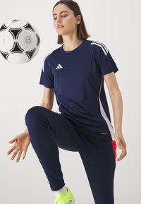 Tumesinine spordipoloshirts valgete triipude ja "AEROREADY" detailidega; tumedad püksid, erksad kollased kingad ja mustvalge jalgpall liikumises.