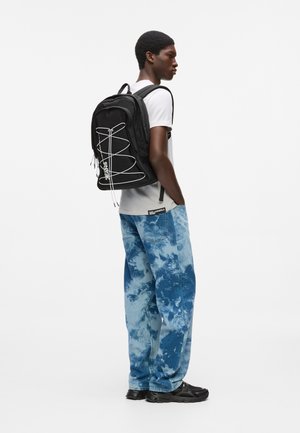 Junger Mann mit schwarzem Rucksack mit weißen Kabelakzenten, Batik-Jeans in Blau, weißem T-Shirt und schwarzen Sneakern, seitlich stehend vor weißem Hintergrund.