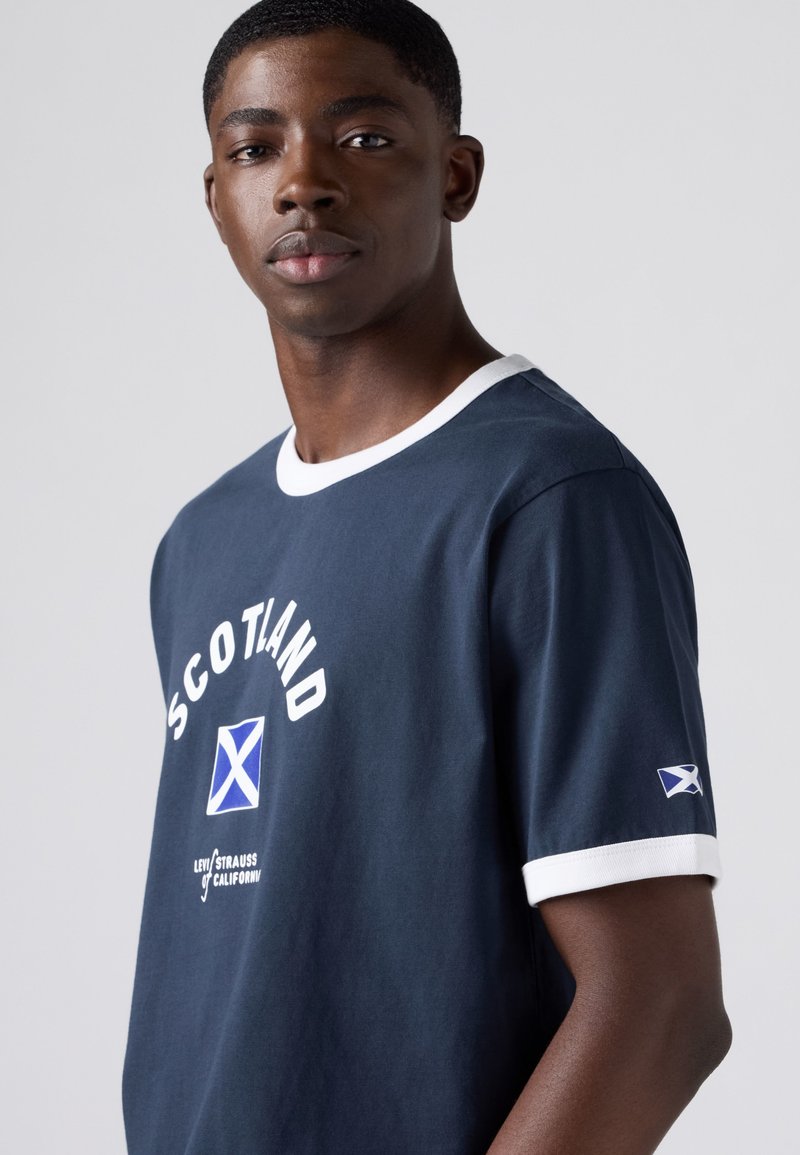 Young man wearing navy blue t-shirt with white collar and sleeve trim, featuring "Scotland" text and Scottish flag on chest and sleeve.