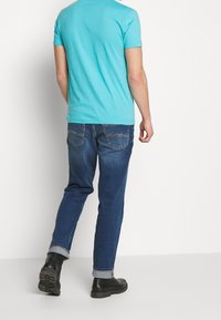 Türkisfarbener Baumwoll-T-Shirt mit kurzen Ärmeln, kombiniert mit dunkelblauen Jeans. Dunkle Stiefel mit strukturierten Sohlen runden das Outfit ab.