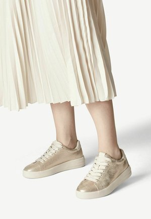 Femme portant des baskets montantes en dentelle métalliques dorées assorties à une jupe midi plissée beige clair sur fond blanc.