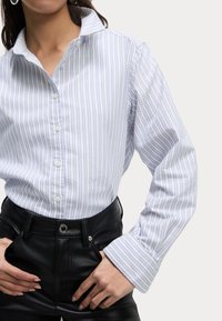 Camisa de botones a rayas azules y blancas con cuello, mangas largas y un corte relajado, combinada con pantalones de cuero negros de cintura alta.