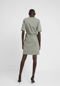 Khaki green dress with short sleeves, a fitted waist, and a straight-cut skirt, made from smooth fabric. The model is wearing black heels.