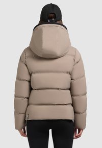 Chaqueta tipo puff en beige claro con capucha, que presenta secciones acolchadas y una textura suave. Combinada con leggings negros y una gorra.