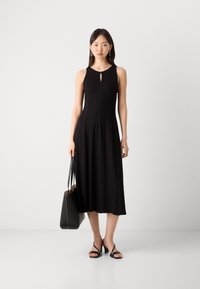 Abito nero senza maniche con scollatura a chiave, corpetto aderente e gonna svasata. Completo di borsa nera e sandali con tacco.
