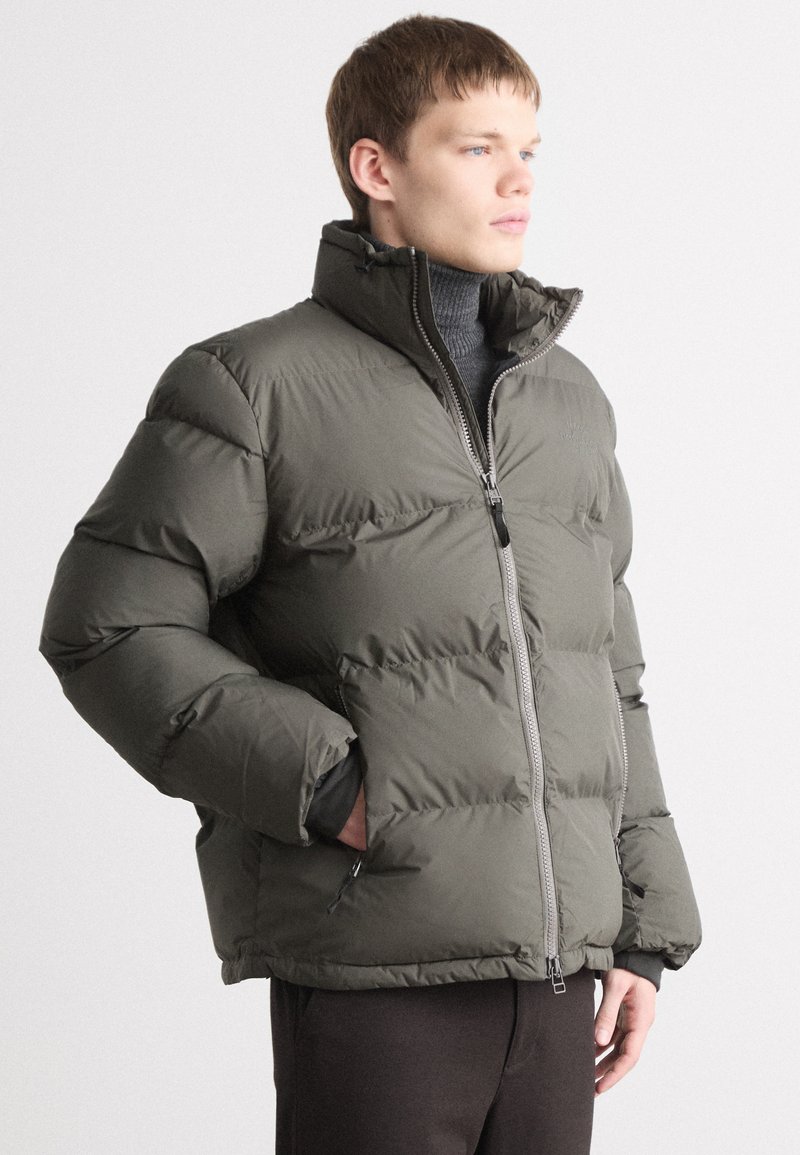 Jachetă puffer de culoare verde măslinie, cu guler înalt, design matlasat, fermoar pe întreaga lungime în față și buzunare laterale. Material texturat cu finisaj mat.
