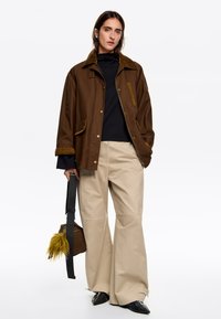 Bruine katoenen jas met een corduroy kraag, gedragen met beige wijde broekspijpen en zwarte puntige schoenen. Model houdt een bruine tas met een veeraccent vast.