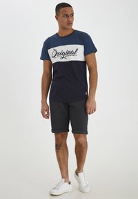 T-shirt bleue marine et blanche avec le texte "Original", associée à un short gris. Style décontracté, manches courtes et poignets retroussés. Baskets blanches.