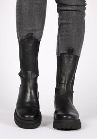 Schwarze Leder-Stiefeletten mit glatter Struktur, elastischen seitlichen Paneelen und einer dicken Gummisohle. Graue Jeans sind teilweise über den Stiefeln sichtbar.
