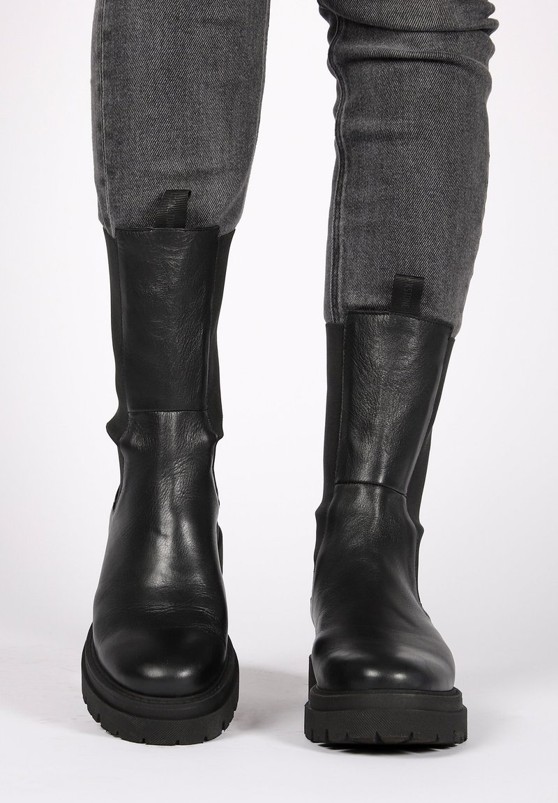 Schwarze Leder-Stiefeletten mit glatter Struktur, elastischen seitlichen Paneelen und einer dicken Gummisohle. Graue Jeans sind teilweise über den Stiefeln sichtbar.
