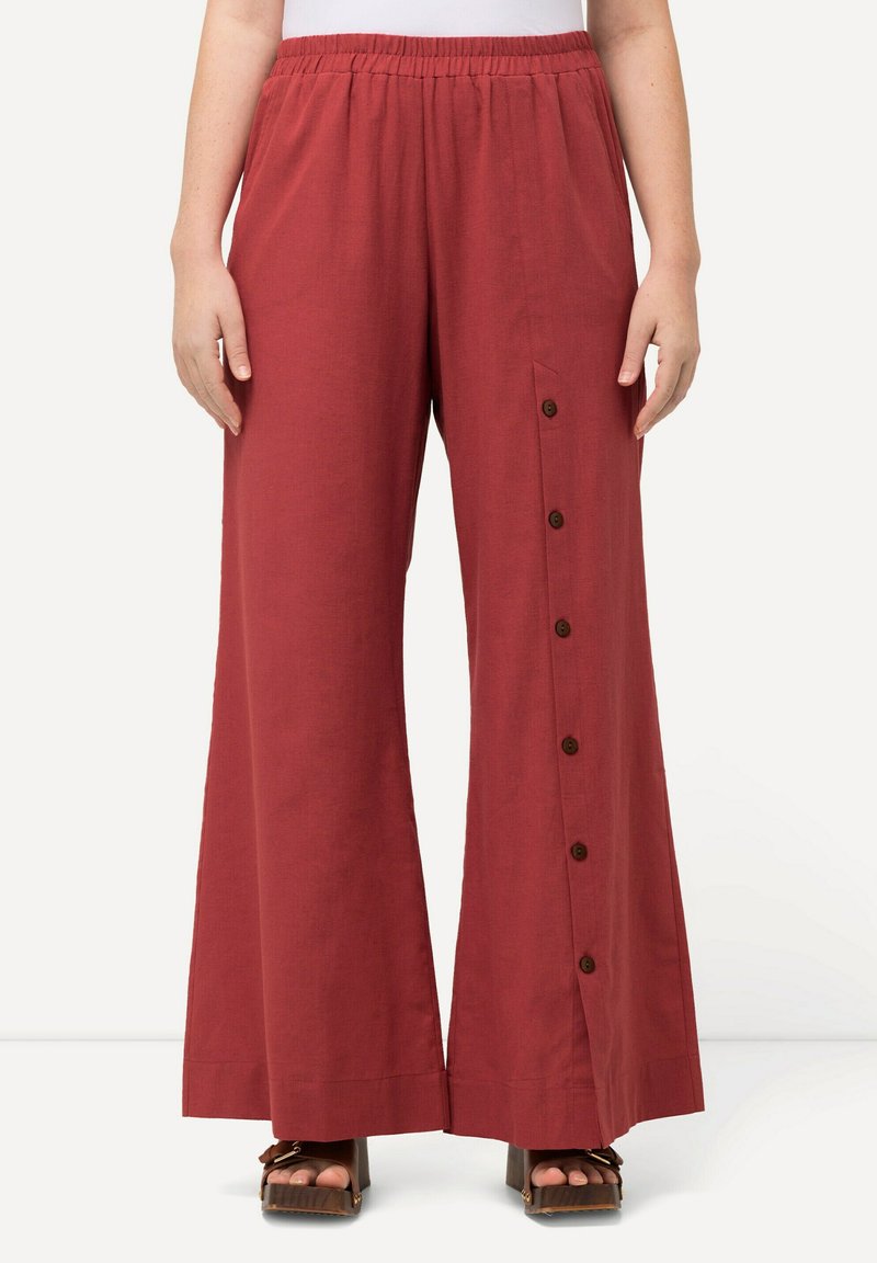 Pantalons larges rouges à taille élastique, avec un détail de bouton sur le côté et un tissu doux et texturé. Portés avec des sandales à plateforme.