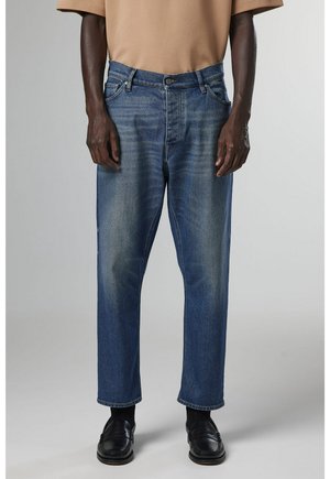 Hombre con vaqueros azules descoloridos, mocasines negros, calcetines negros y una camisa de manga corta beige, de pie frente a un fondo liso.