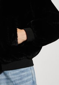 Sort faux fur jakke med en blød tekstur og ribbede sorte manchetter, stylet med lyseblå denim. Designet fremhæver komfort og varme.