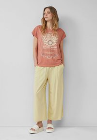 Rustkleurig T-shirt met korte mouwen en een zonnenafbeelding, gecombineerd met lichtgele wijde broek en witte platformsandalen.