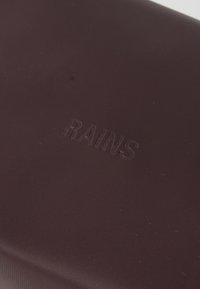 Borsa di un rosso vinaccia scuro con una finitura liscia e opaca. Presenta il logo "RAINS" in rilievo. Forma rettangolare semplice, superficie testurizzata.