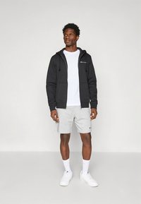 Zwarte capuchontrui met rits en een logo, wit T-shirt eronder, grijze korte broek en witte sneakers. De outfit heeft een casual, sportieve uitstraling.