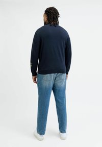Navy knit sweater with a crew neck, long sleeves, and colorful patch on the left sleeve. Paired with light blue straight-leg jeans and white sneakers.