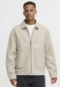 Beige uldblandingsjakke med lynlås fortil, store påsyede lommer og spidse kraver. Båret over en hvid skjorte, med mørke jeans.