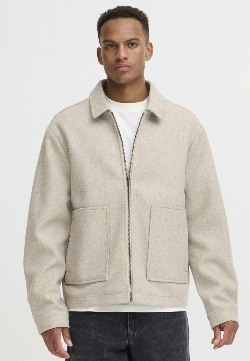 Beige uldblandingsjakke med lynlås fortil, store påsyede lommer og spidse kraver. Båret over en hvid skjorte, med mørke jeans.