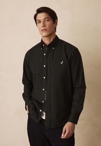 Tmavozelená košeľa na gombíky z textúrovanej látky s bielymi gombíkmi, s vyšívaným logom vtáka na ľavej strane hrudníka.