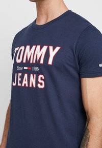 Navy blue cotton t-shirt featuring bold white and red "TOMMY JEANS" text. Short sleeves and a crew neckline. Simple and casual design.