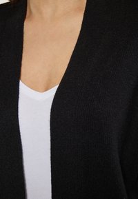 Cardigan negru cu o textură moale și un design cu față deschisă, asortat cu un maiou alb cu guler în V, evidențiind culori contrastante.
